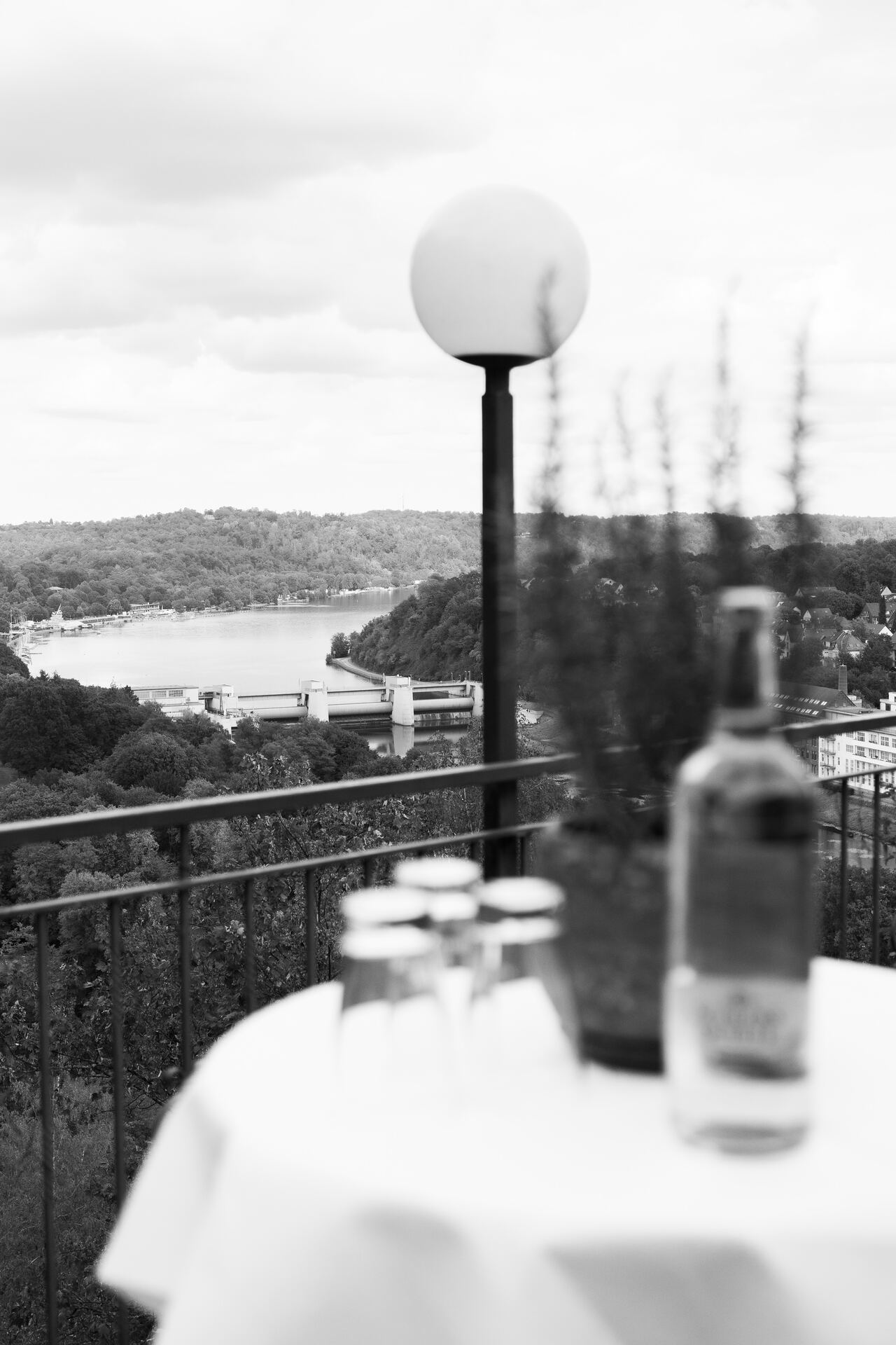 Stehtisch mit Flaschen auf dem Balkon, dahinter der Blick über den See, Schwarzweiß